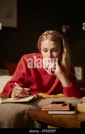 Vertical image of young Caucasian woman lying on bed and writing in notebook, planning schedule. Image represents mental wellbeing and is suitable for lifestyle blog marketing Stock Photo