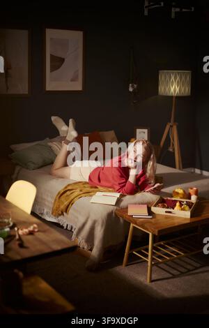Vertical image of thoughtful Caucasian young woman lying on bed, holding smartphone and listening to music in headphones, perfect for mental wellbeing and cozy lifestyle campaigns Stock Photo