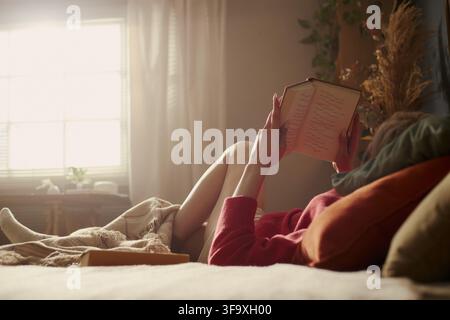 Young Caucasian woman lying on bed and reading book, relaxing at home. Image represents mental wellbeing and self care, perfect for lifestyle and leisure marketing campaigns Stock Photo