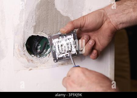 Electrician is using a screwdriver to install an electrical outlet on a white wall. The surroundings are minimalistic, indicating a recent home renova Stock Photo