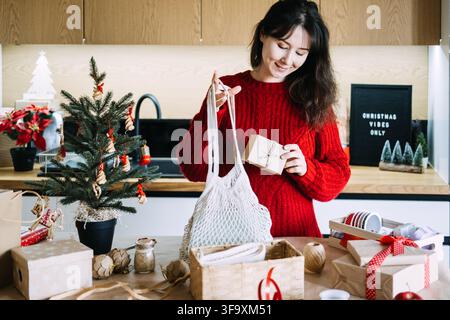 Woman holds mesh bag and kraft wrapped parcel in kitchen near CHRISTMAS VIBES ONLY sign. Eco christmas gift business, sustainable gift shop christmas Stock Photo