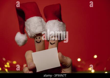 Fingers dressed in Santa-Claus red-white hats. Two happy finger smileys faces holding empty paper note for your text copy space against red background Stock Photo