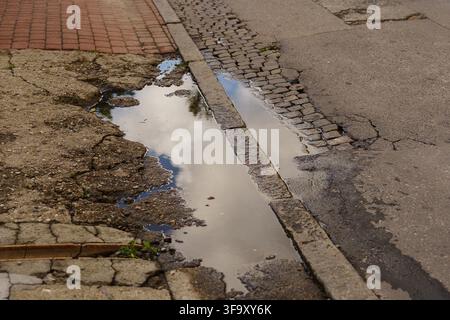 Rain puddle on a cracked, weathered asphalt street, reflecting a cloudy sky, illustrating urban infrastructure decay and road surface texture Stock Photo