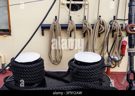 A detailed shot of a ship's deck featuring thick black ropes coiled around twin bitts. In the background, several rigging ropes hang neatly coiled on Stock Photo
