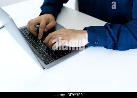 Close-up of a businessman in a dark blue shirt actively typing on a modern laptop keyboard at a clean white office desk. Stock Photo