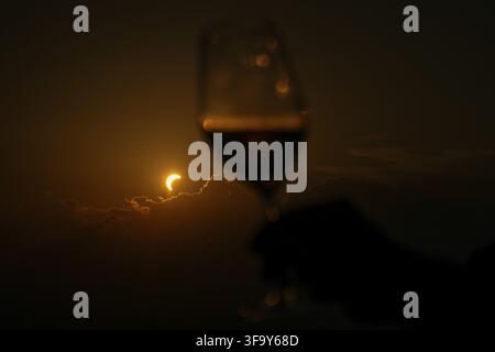 View of a partial solar eclipse seen through a wine glass held by a hand against a dark, cloudy sky in Turin, Piedmont, Italy. Stock Photo