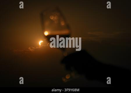 View of a partial solar eclipse seen through a wine glass held by a hand against a dark, cloudy sky in Turin, Piedmont, Italy. Stock Photo