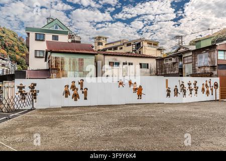 An unbroken row of wooden silhouettes – women, spear bearers and children in historical dress – stretches along a construction hoarding on Hayakawa-dori (早川通り) in Hakone-Yumoto, Kanagawa Prefecture, Japan, in front of old wooden houses and shops. The imagery likely evokes the Hakone Daimyo Gyoretsu, an annual procession held on this very street every 3 November since 1935. Stock Photo