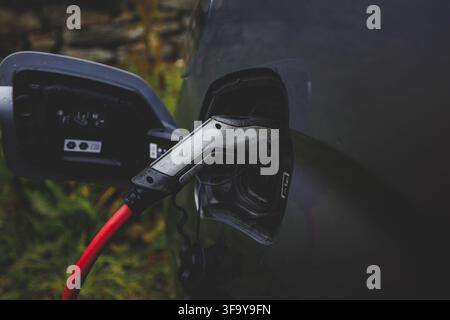 Close-up of a charging plug connected to a red EV in rainy Norway in late summer, symbolizing electrification and humanity’s move beyond fossil fuels Stock Photo