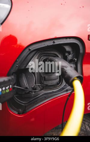 Close-up of a charging plug connected to a red EV in rainy Norway in late summer, symbolizing electrification and humanity’s move beyond fossil fuels Stock Photo