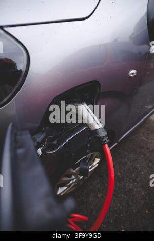 Close-up of a charging plug connected to a red EV in rainy Norway in late summer, symbolizing electrification and humanity’s move beyond fossil fuels Stock Photo