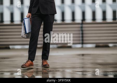 Businessperson walking in suit with leather shoes and documents reflected on wet pavement Stock Photo