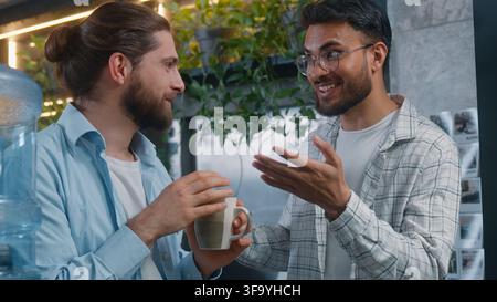 Two multiracial men Caucasian Indian male friends talking laughing in office cafe startup project casual guys business colleagues talk happy laugh Stock Photo