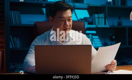 Happy motivated middle-aged Korean business man working laptop paperwork in night office Chinese businessman typing computer managing papers documents Stock Photo