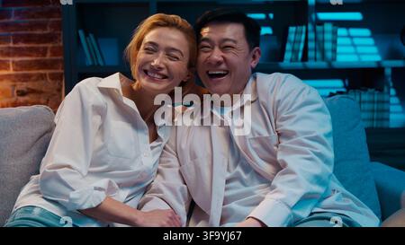 Happy cheerful multiracial couple watching television laughing enjoying spending time together at evening living room multiethnic spouses Asian man Stock Photo