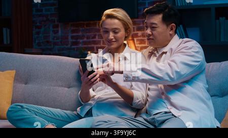 Multiethnic couple emotional discussing shopping using mobile phone at evening living room at couch. Multiracial family Asian man with Caucasian woman Stock Photo