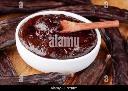 thick carob spread with a glossy chocolate-like texture fills a white bowl, with a wooden spoon resting on top and dried carob pods scattered around o Stock Photo