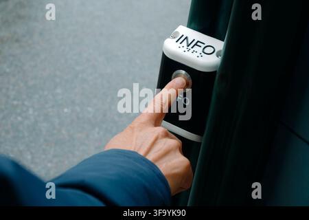 a man presses a silver button on an outdoor public information device mounted on a dark green metal post, with braille lettering above and pavement so Stock Photo