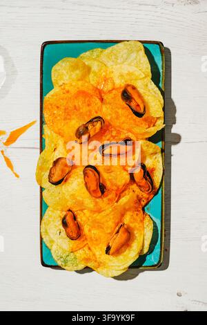 seen from above, mussels and potato chips coated with escabeche sauce, a popular Spanish bar or home aperitif, fill a turquoise plate on a light woode Stock Photo