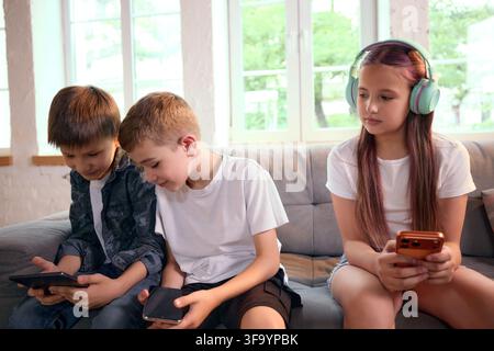 Three school-age friends using personal mobile devices together on sofa Stock Photo