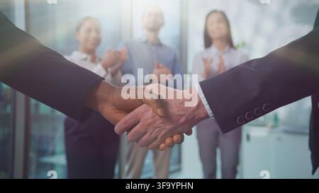 Two diversity men executives shaking hands in sun business office near colleagues applauding. Multiethnic businessmen shake arms team concluding Stock Photo