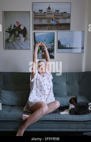 Happy young woman stretching her arms while sitting on sofa at home with her pet cat. Stock Photo