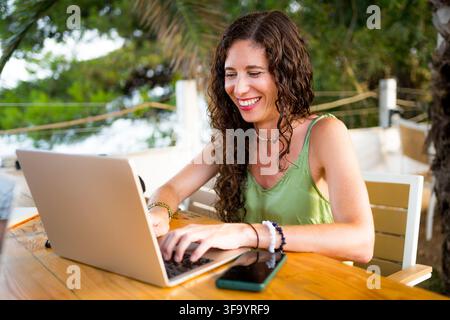 smiling young woman doing remote work on notebook at outdoor terrace cafe bar, happy millennial in her 40s digital nomad lifestyle Stock Photo
