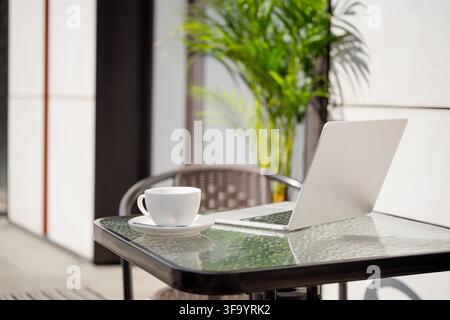 Laptop and white coffee cup on a sunny outdoor cafe table surrounded by green plants, modern remote workplace with nobody and copy space. Stock Photo