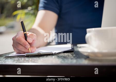 Close up of Caucasian man writing notes with pen beside laptop and coffee at outdoor cafe, remote work, planning and productivity concept. Stock Photo
