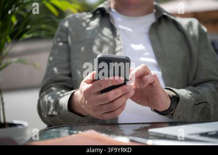 Close up of a Caucasian man using smartphone at an outdoor cafe table with laptop, modern mobile technology and remote work concept. Stock Photo