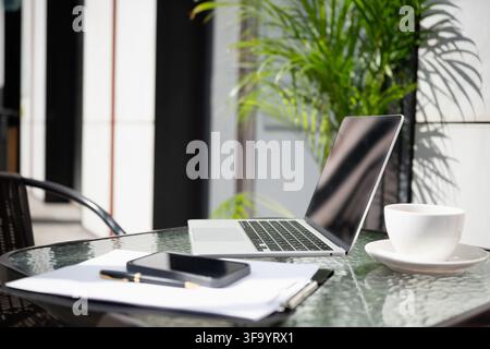 Laptop, smartphone, notebook and coffee arranged on sunny outdoor cafe table, modern freelance workplace and digital nomad lifestyle with nobody. Stock Photo