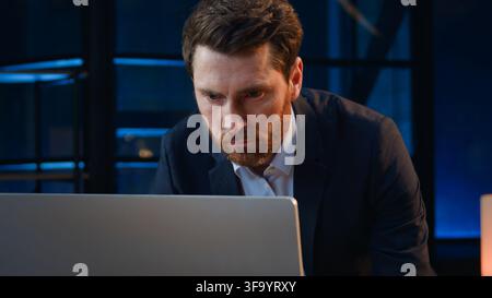 Caucasian serious focused middle-aged Caucasian business man looking in computer at dark night evening office mature businessman male executive Stock Photo