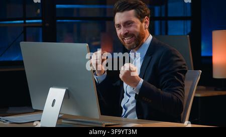 Successful happy Caucasian mature business man winning with computer dancing at dark evening office at late night cheerful positive middle-aged Stock Photo