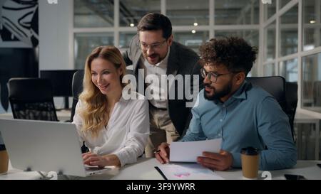 Three happy excited multiethnic business workers working with laptop documents talking in office boss rejoice good work diversity managers excellent Stock Photo