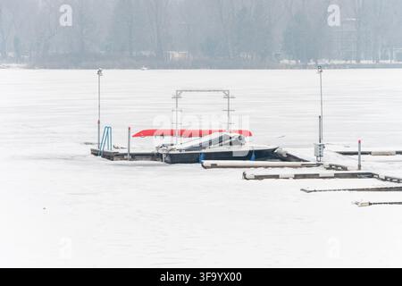 A covered motorboat sits at a snow-dusted dock on a frozen misty lake. Stock Photo