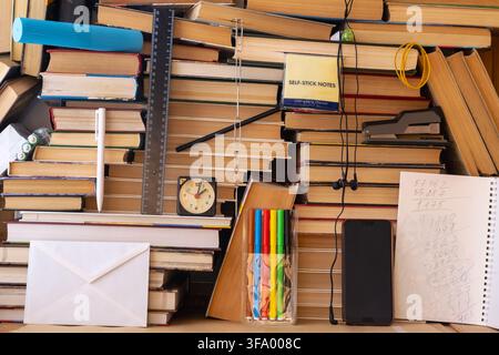 A cluttered study workspace made from stacked books holds pens, colored markers, ruler, clock, sticky notes, smartphone, earbuds and a notepad. Stock Photo