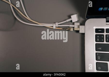 Laptop charger cable with frayed, exposed wires and damaged insulation plugged into a laptop keyboard, highlighting unsafe power connection, wear and Stock Photo