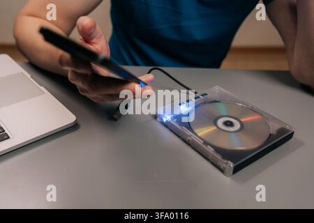 Male hand connects smartphone to external optical disc drive on a desk beside laptop, showing a media disc and glowing blue usb ports for data Stock Photo