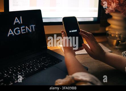 Individual interacts with smartphone showing AI AGENT text at a desk, with a laptop and computer screen displaying data charts in the background Stock Photo