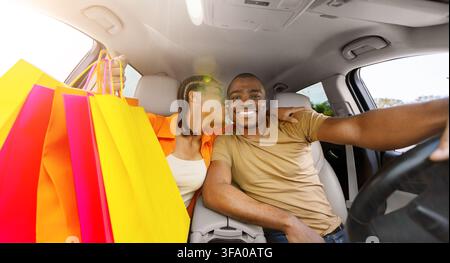 Black woman holding colorful shopping bags while kissing her partner inside car Stock Photo