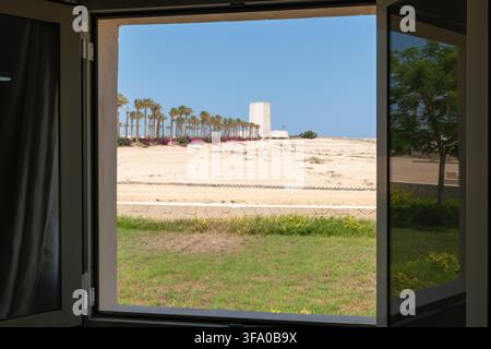 View of the Italian Cemetery at Al Alamein War Memorial with palm trees and desert landscape under a clear blue sky, seen through a window in Al Alame Stock Photo