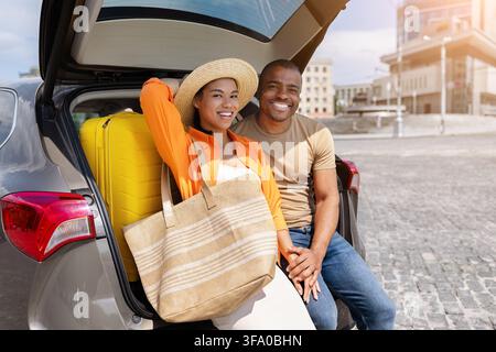 Smiling black couple sitting in car trunk with suitcase and beach bag Stock Photo