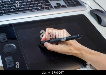 Designer's hand draws with a digital stylus on a black graphics tablet on a desktop next to a computer keyboard. Creative process of digital illustrat Stock Photo