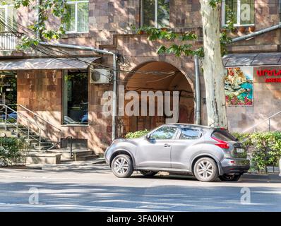 Silver SUV parked near stone archway building. Tree branches cast shadows on street. Greenery lines sidewalk beside historic architecture. No animals Stock Photo