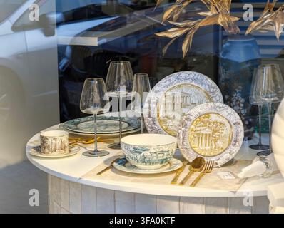 Elegant dinnerware set on white round table. Plates, bowls, glasses, and cutlery arranged. Decorative gold leaf accents on plates. Modern glassware an Stock Photo