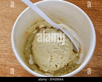 Vanilla ice cream with chocolate chips in white cup. Plastic spoon rests inside. Wooden surface beneath. Slightly melted texture visible Stock Photo