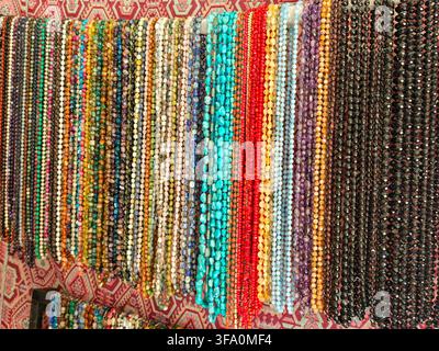 Vibrant beads hang in rows, colorful and varied. Some beads are turquoise, red, gold, black. They are strung on threads. Beads are displayed against p Stock Photo