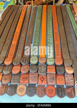 Rows of leather belts with intricate embossed patterns and metal buckles. Belts vary in color from brown to green and orange. Each belt features uniqu Stock Photo