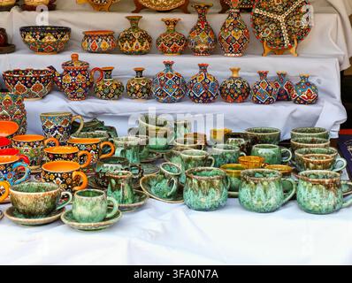 Colorful ceramic pots and cups arranged on white display tables. Vessels feature intricate painted patterns in red, green, yellow, and brown. Some cup Stock Photo