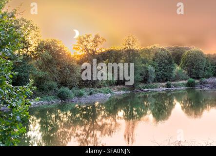 An 80 percent partial solar eclipse glowing in the sky over a calm lake and dense forest background during golden hour daylight. Breathtaking astronom Stock Photo
